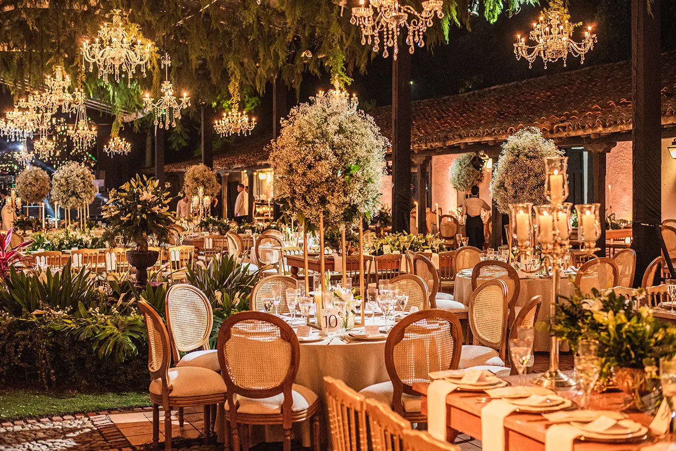 Banquet with chandeliers, wooden chairs and flowers under lit trees